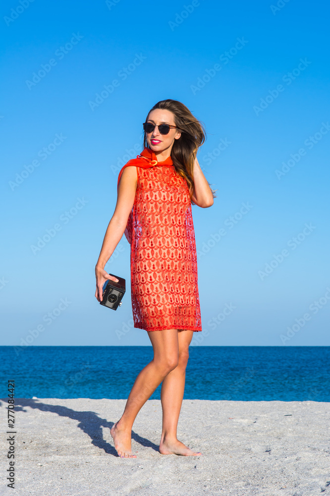 Retro fashion look. Pretty cool young woman model with retro film camera wearing an elegant red colored vintage dress (60ies style), blonde hair with sunglasses at the beach.