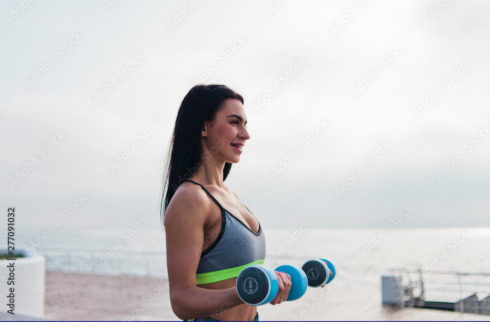 Young fit woman in sportswear doing exercise with dumbbells at the morning on sunrise beach Young fit woman in sportswear doing exercise with dumbbells at the morning on sunrise beach