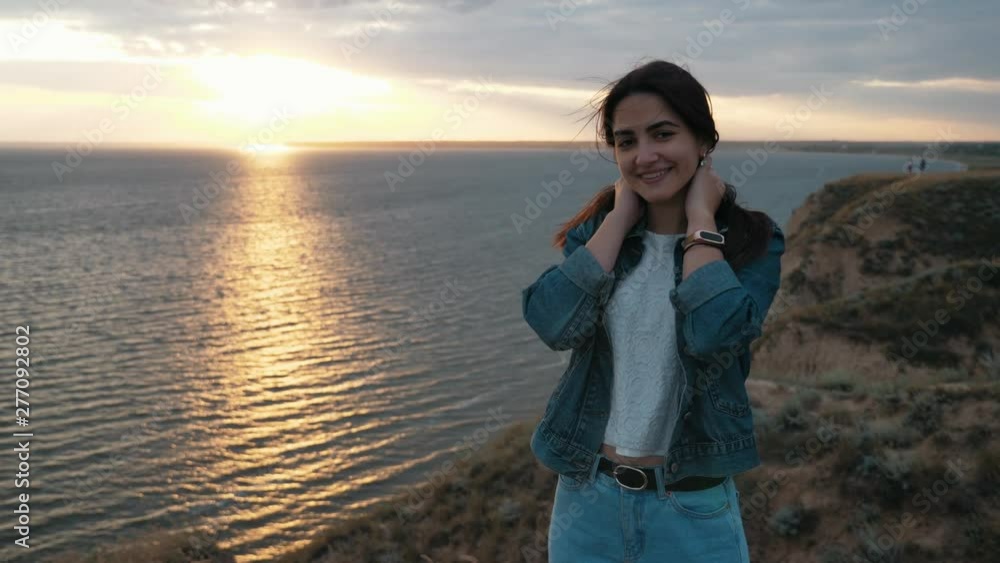 Positive awesome brunette girl in jeans jacket standing on the mountain near the sea with strong wing waving her hair at sunset, Slow motion