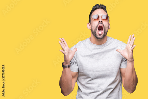 Young handsome man wearing sunglasses over isolated background crazy and mad shouting and yelling with aggressive expression and arms raised. Frustration concept.