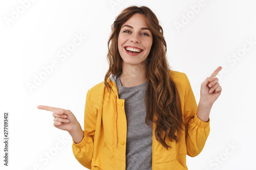 Enthusiastic silly curly-haired beautiful young girl curly stylish hairstyle pointing sideways index fingers left right smiling delighted pleased lots choices good offers, standing white background photo