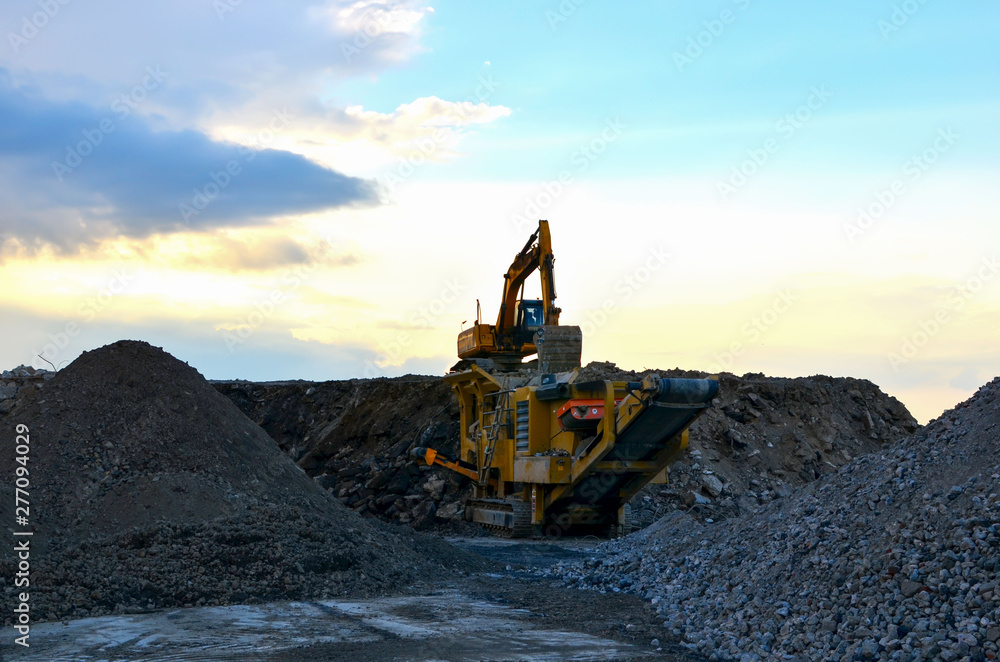Mobile Stone crusher machine by the construction site or mining quarry for crushing old concrete slabs into gravel and subsequent cement production. Heavy excavator on a background of sunset