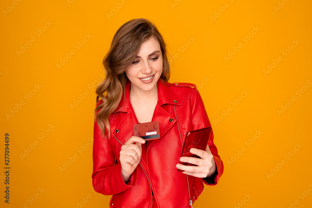 Naklejka premium Pretty girl in red clothes holding phone and credit card isolated over the bright yellow background