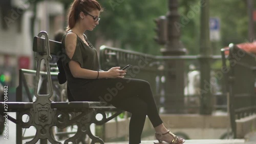Attractive caucasian impatient woman with glasses, freckles, piercings and red hair waiting and watching at her smartphone sitting on street bench, during sunny summer in Paris. Slow motion.