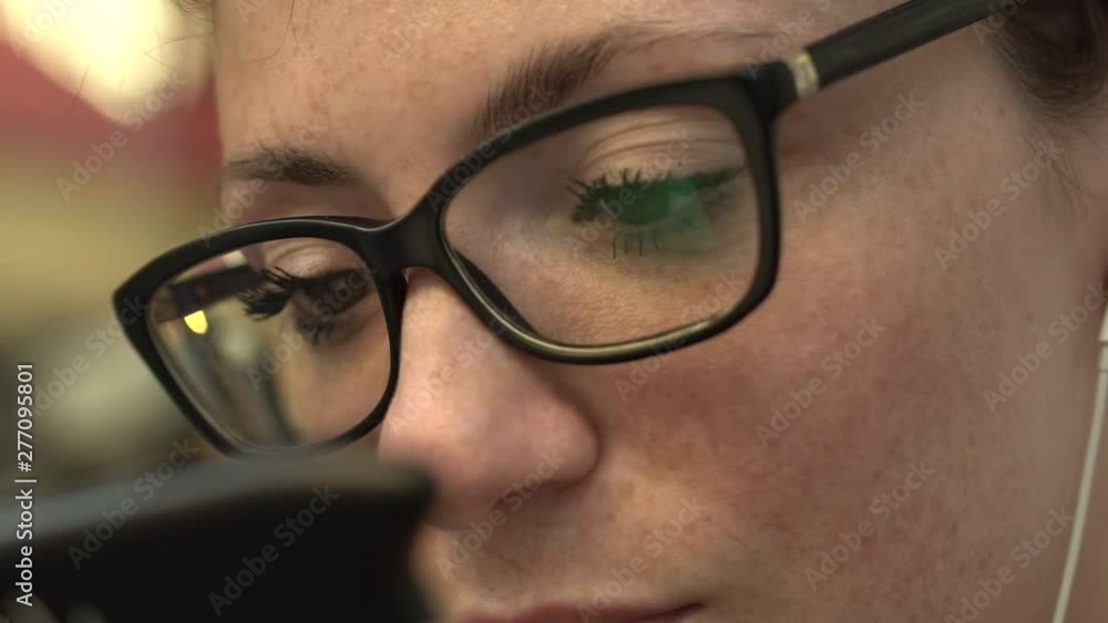 Close up of eyes of attractive smiling redhead woman with glasses reflection, freckles and red hair watching internet video on smartphone sitting at bus stop in street in Paris. Flashing green light. Close up of eyes of attractive smiling redhead woman with glasses reflection, freckles and red hair watching internet video on smartphone sitting at bus stop in street in Paris. Flashing green light.