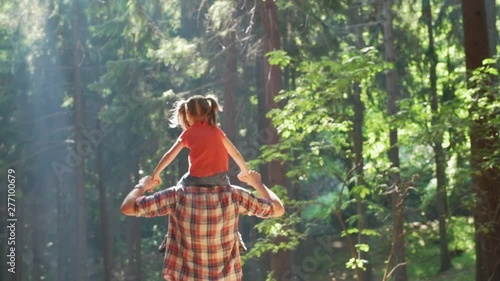 Back view of father giving a piggyback ride to daughter walking in forest.Father holding daughter on shoulders hiking along foresth trail path.Father and daughter hiking on forest path together