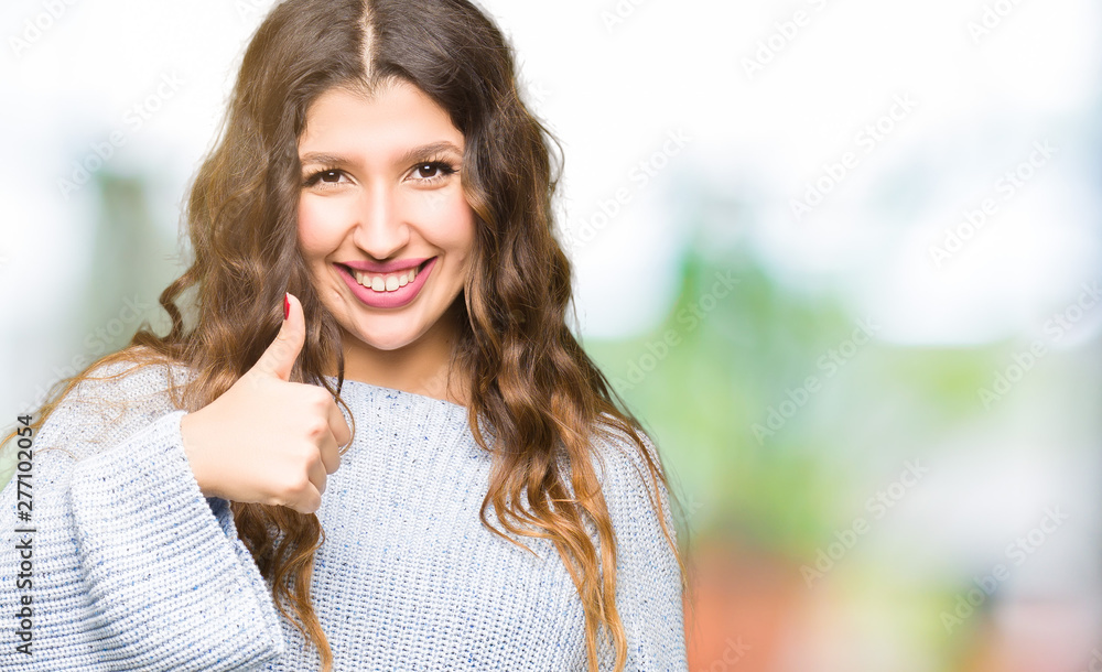 Young beautiful woman wearing winter sweater doing happy thumbs up gesture with hand. Approving expression looking at the camera showing success.