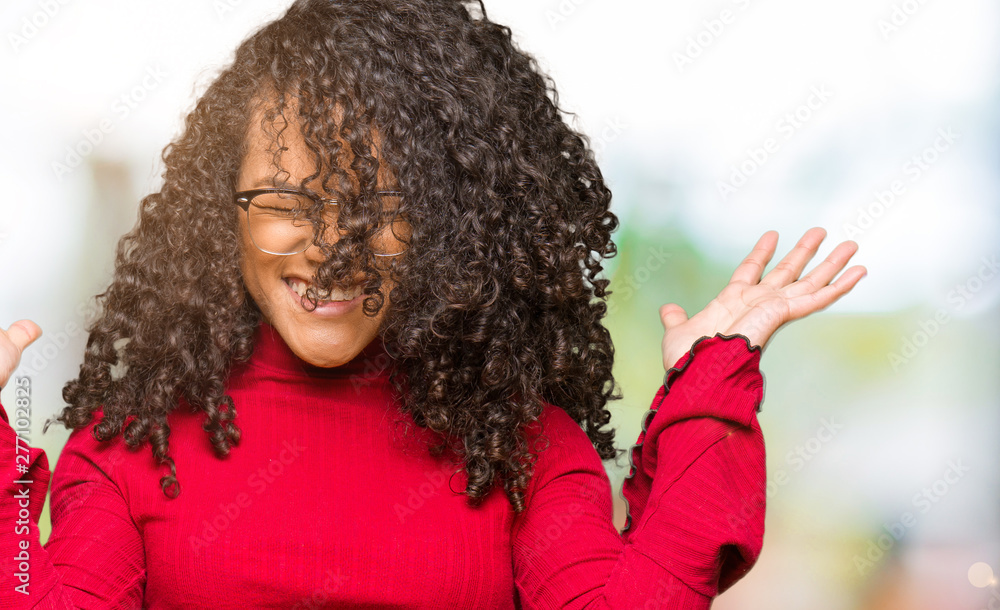 Young beautiful woman with curly hair wearing glasses celebrating mad and crazy for success with arms raised and closed eyes screaming excited. Winner concept Young beautiful woman with curly hair wearing glasses celebrating mad and crazy for success with arms raised and closed eyes screaming excited. Winner concept