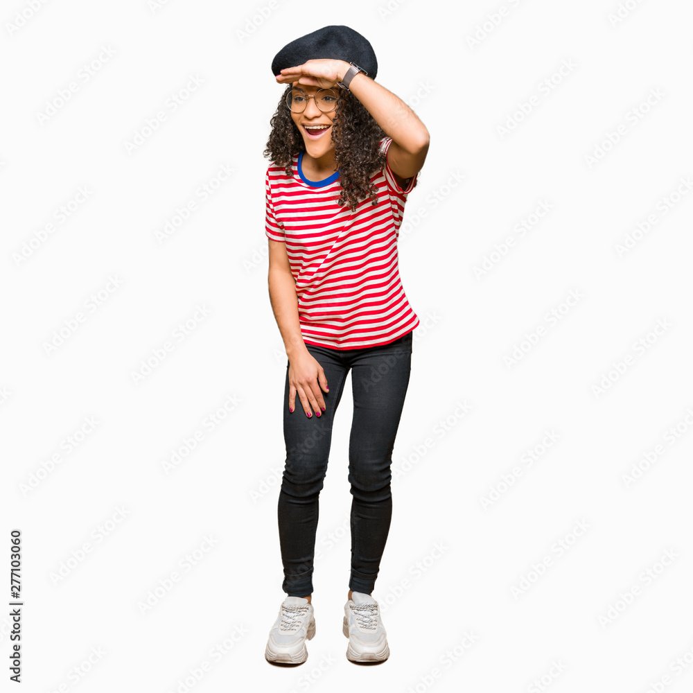 Young beautiful woman with curly hair wearing glasses and fashion beret very happy and smiling looking far away with hand over head. Searching concept. Young beautiful woman with curly hair wearing glasses and fashion beret very happy and smiling looking far away with hand over head. Searching concept.