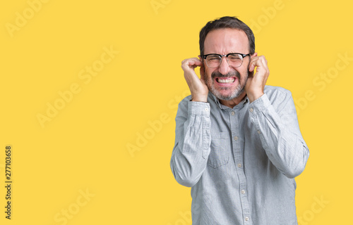 Handsome middle age elegant senior man wearing glasses over isolated background covering ears with fingers with annoyed expression for the noise of loud music. Deaf concept. © Krakenimages.com