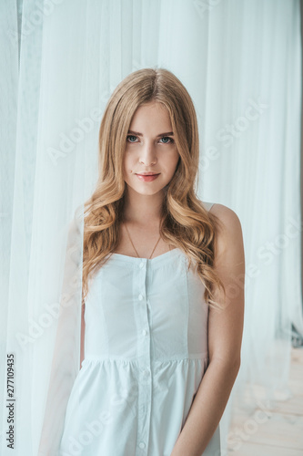 Girl in a light white dress posing by the window.