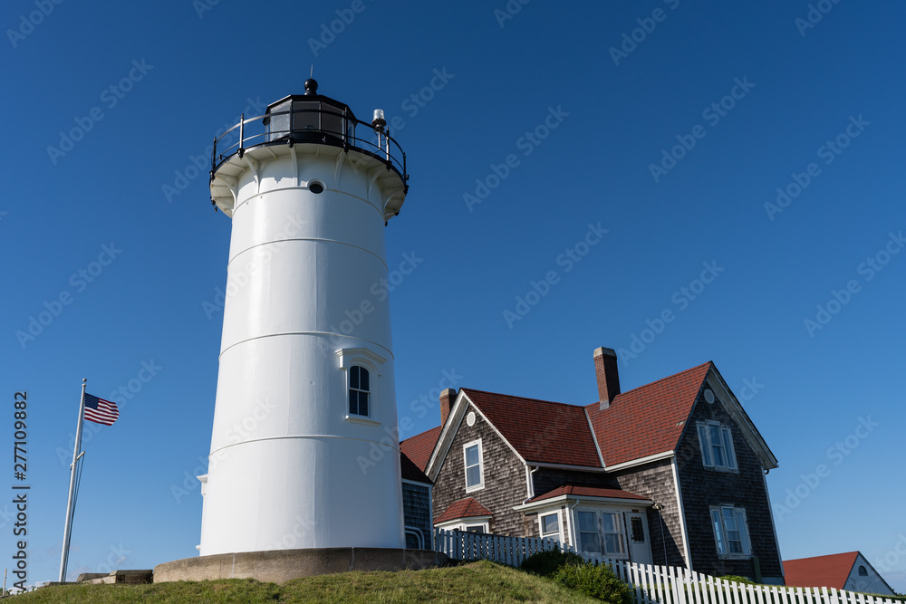 Nobska Lighthouse with a bright blue sky and the red roofed house beside it and a white fence and the USA flag