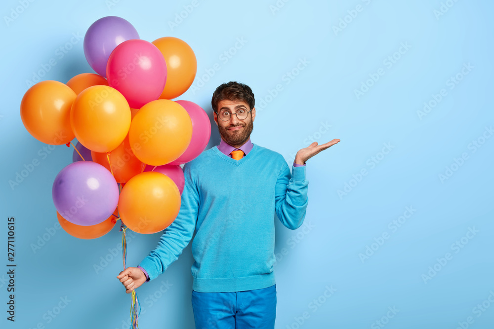 Confused hesitant male holiday maker raises palm with puzzlement, tries to make decision, holds multicolored bunch of air balloons, doesnt know whether all guests has come, wears blue clothes - obrazy, fototapety, plakaty 