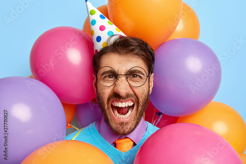 Spoiled holiday concept. Annoyed dissatisfied man shouts and frowns face in discontent, wears cone hat, has office party, stands around inflated colorful balloons, expresses negative emotions