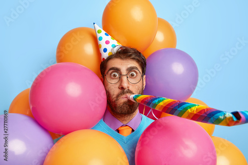 Good looking man has surprised face, blows into party horn, congratulates daughter with birthday, uses holiday attributes, poses against colorful balloons, enjoys celebration, shocked by something