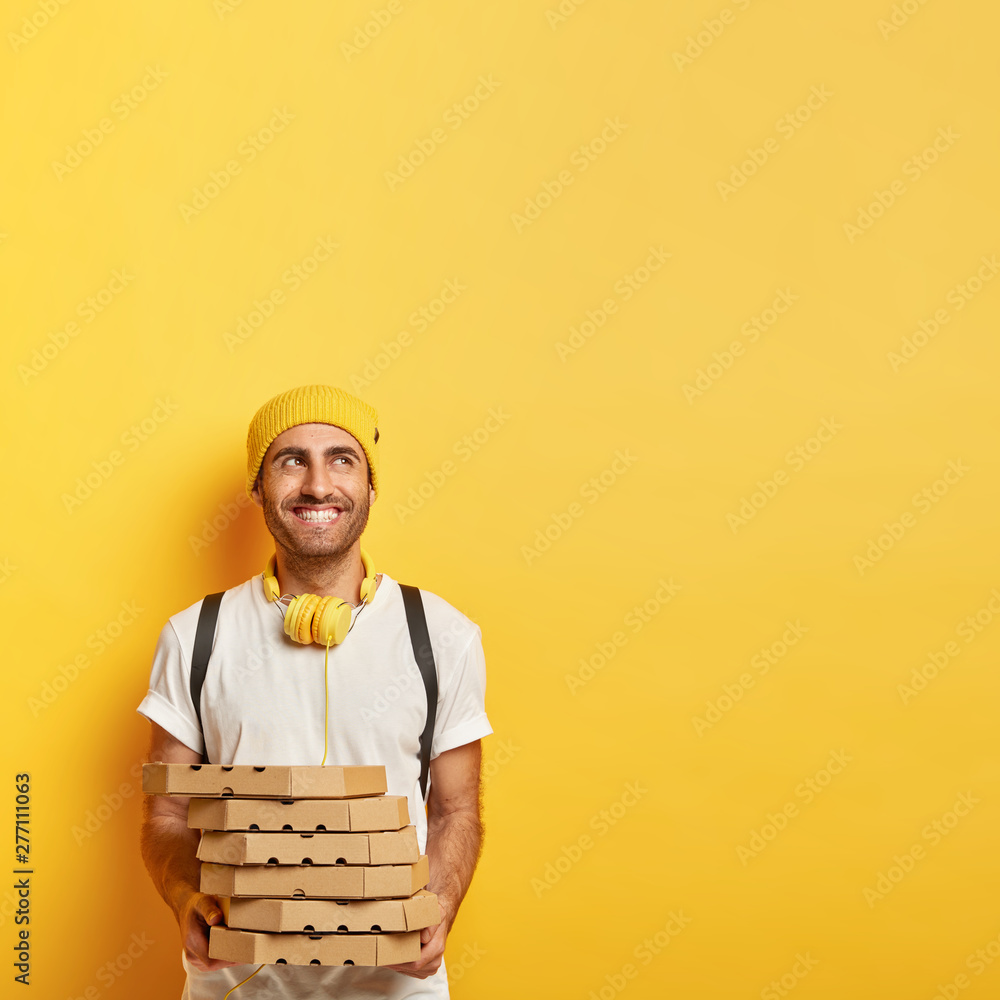 Positive delighted pizza man holds stuck of cardboard containers, waits for payment, does duties, looks aside, has broad smile, wears stylish outfit, isolated on yellow wall, copy space for promotion Positive delighted pizza man holds stuck of cardboard containers, waits for payment, does duties, looks aside, has broad smile, wears stylish outfit, isolated on yellow wall, copy space for promotion