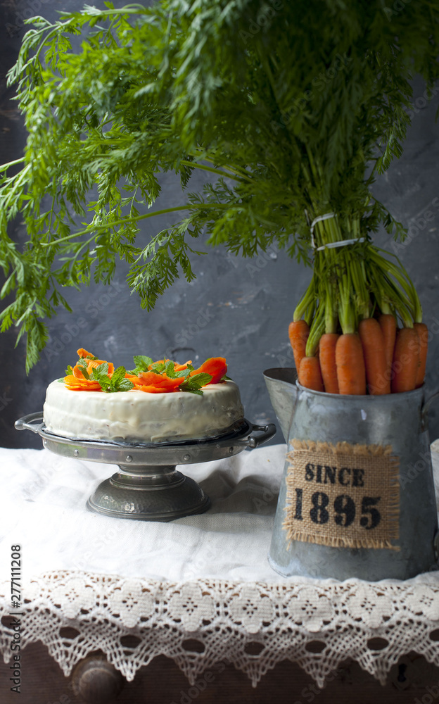 Carrot cake pie, sprinkled with nuts, decorated with cream-colored carrots on a stand for cakes, fresh carrots in a white box on a dark black background wooden table in rustic style