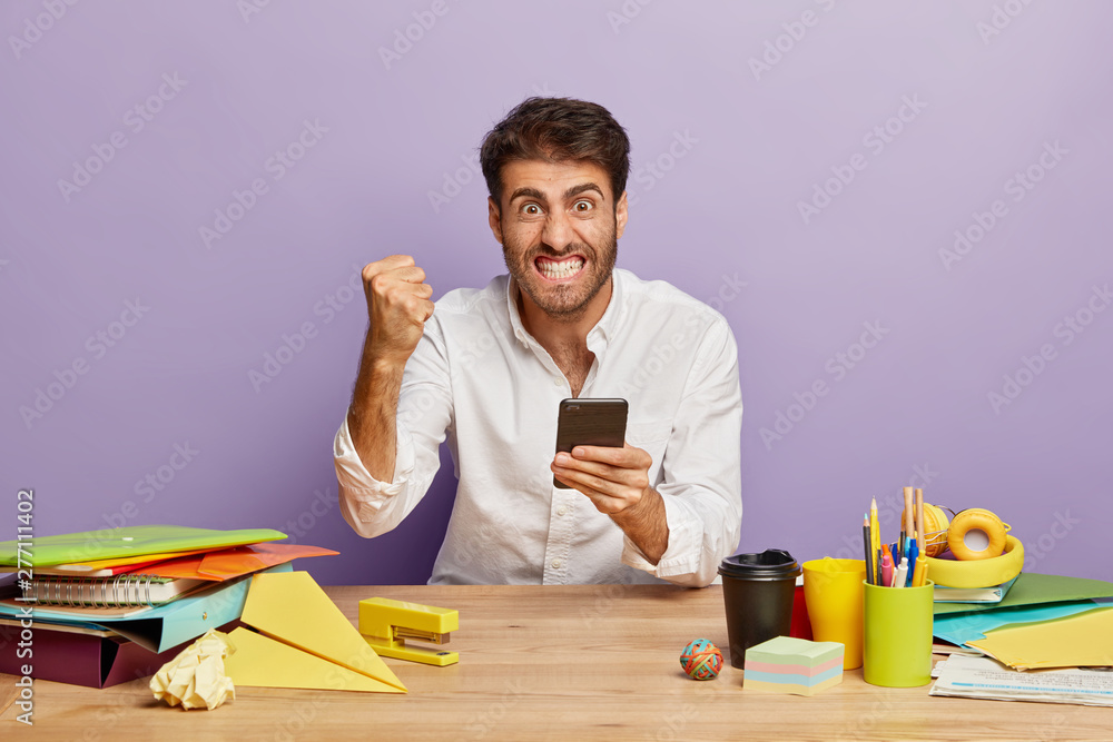 Horizontal shot of irritated student clenches teeth and fist with anger, holds modern cell phone in hands, receives notification to pay bills, wears white shirt, sits at wooden desk stationery around Horizontal shot of irritated student clenches teeth and fist with anger, holds modern cell phone in hands, receives notification to pay bills, wears white shirt, sits at wooden desk stationery around