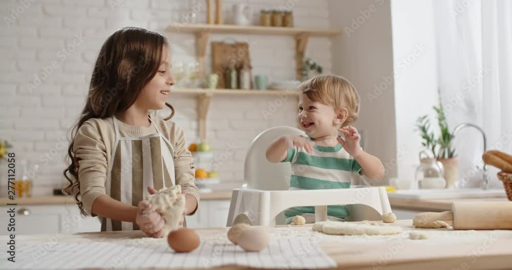 Little girl is learning to cook, preparing cake dough while her little brother in baby chair with floured face is playing with piece of pastry - togetherness, childhood dream 4k Little girl is learning to cook, preparing cake dough while her little brother in baby chair with floured face is playing with piece of pastry - togetherness, childhood dream 4k