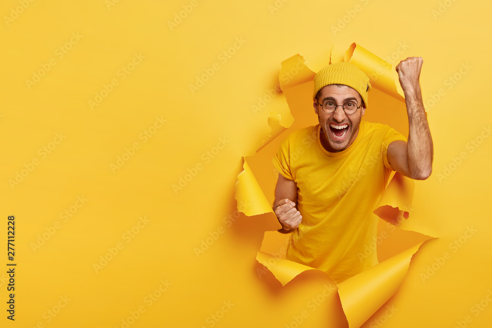 Joyous cheering guy clenches fists with triumph, happy win game, has successful day, wears yellow outfit, stands inside of torn paper hole, free space on left for your text information. Nice work