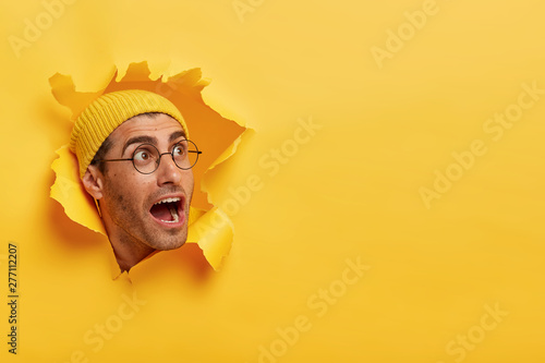 Overwhelmed young guy keeps head through torn hole in yellow background, keeps jaw dropped, gazes aside on free space, notices something surprising, wears hat and big round glasses. Reaction
