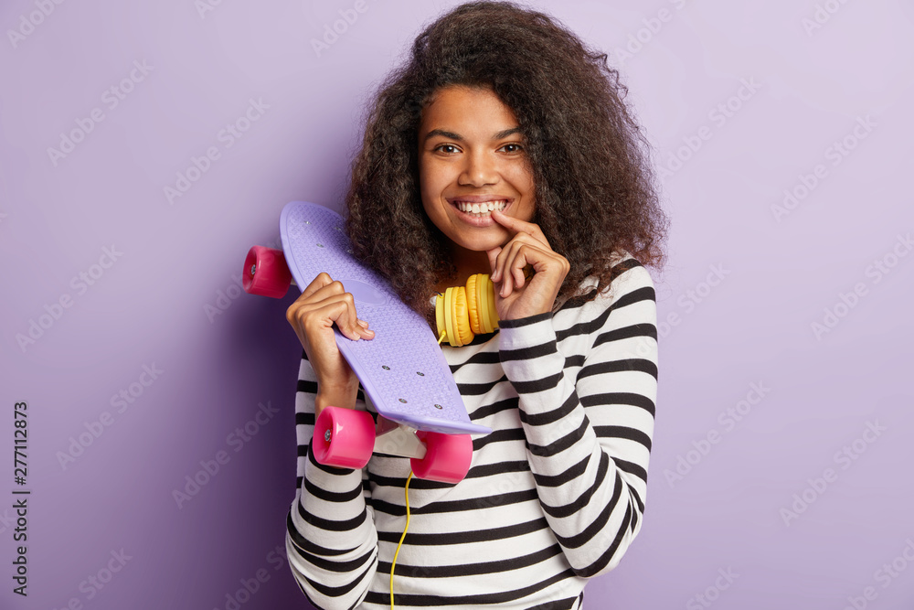 Brunette charming woman has tender smile, keeps index finger on lip, holds purple skateboard, feels free, wears striped jumper, enjoys hobby, isolated purple background, being on way to skate park Brunette charming woman has tender smile, keeps index finger on lip, holds purple skateboard, feels free, wears striped jumper, enjoys hobby, isolated purple background, being on way to skate park
