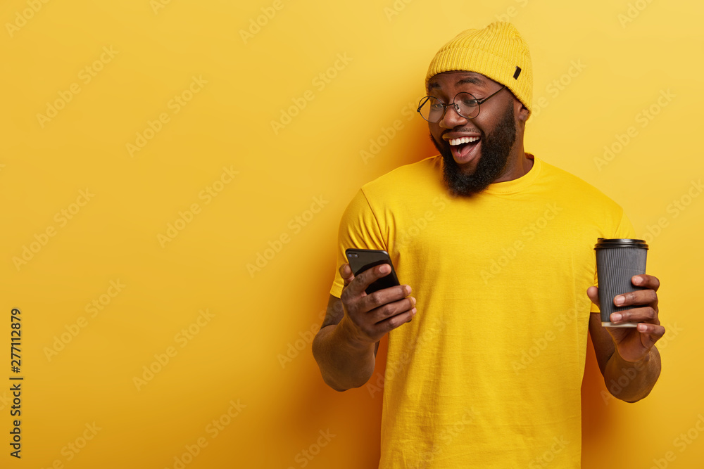 Cheerful dark skinned man uses smartphone application, watches video in internet, chats online, drinks coffee from disposable cup, wears yellow clothes, stands indoor, ignores live communication