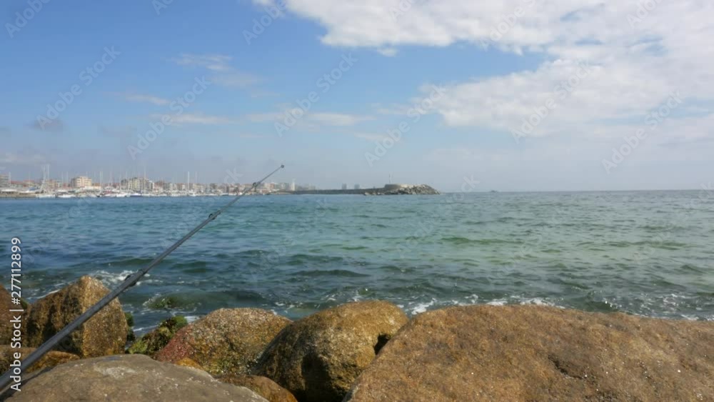 Fishing rod rests on rocks at entrance to harbor as waves break on shore. Bright summer day with blue sky. Fishing rod rests on rocks at entrance to harbor as waves break on shore. Bright summer day with blue sky.