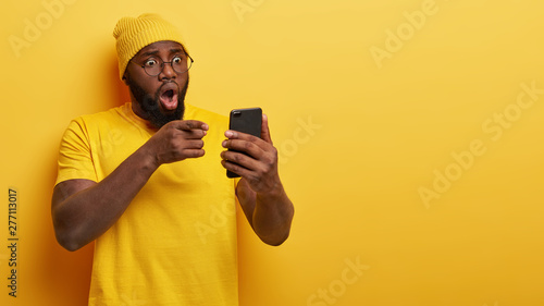Canvas Print Nervous worried dark skinned hipster points at screen, keeps mouth wide opened, sees terrified message content, holds modern mobile phone, wears yellow outfit in one tone with background