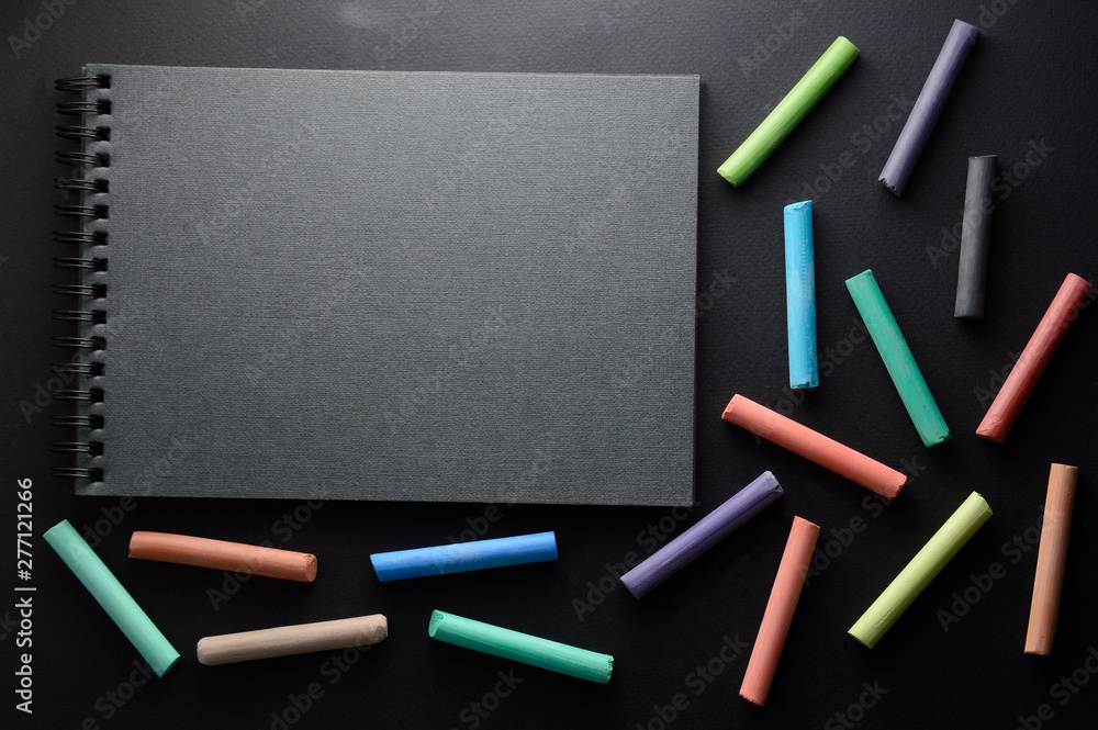 Bright multi-colored chalk on a black background. Sketchbook for drawing pencils or chalk lies on a dark background Bright multi-colored chalk on a black background. Sketchbook for drawing pencils or chalk lies on a dark background
