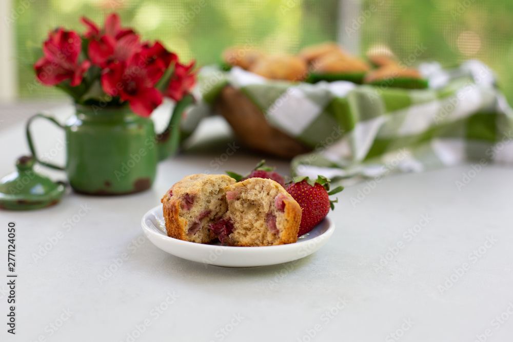 Freshly Baked Strawberry Muffins in wooden bowl with one isolated in front on plate; green and white checked cloth lining bowl; red flowers in green tin. Freshly Baked Strawberry Muffins in wooden bowl with one isolated in front on plate; green and white checked cloth lining bowl; red flowers in green tin.