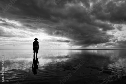 Fotografi Moody monochrome silhouette of man in hat standing in calm shallow waters reflec
