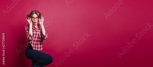 Inspired barefooted girl in glasses standing on one leg in studio. Indoor photo of enthusiastic young woman in checkered shirt posing on claret background.