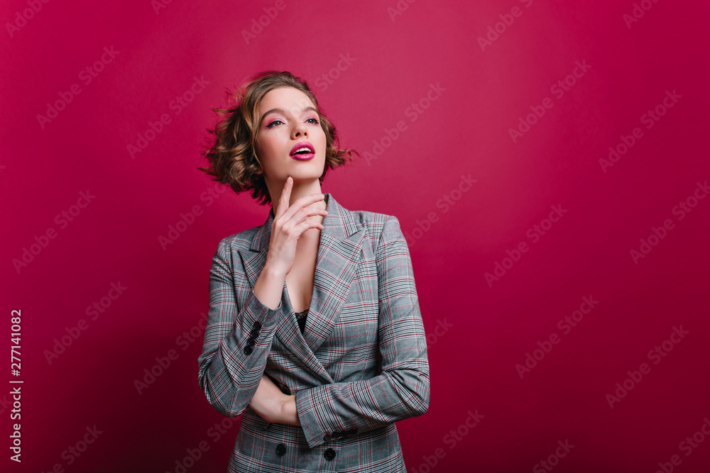 Beautiful confident businesslady posing on claret background and looking away. Dreamy caucasian girl with short haircut in gray jacket standing in studio. Beautiful confident businesslady posing on claret background and looking away. Dreamy caucasian girl with short haircut in gray jacket standing in studio.
