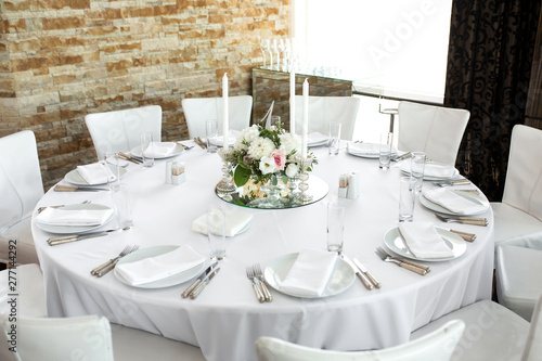 Wedding table setting decorated with fresh flowers. White plates, silverware, white tablecloth and white room. Wedding floristry. Banquet table for guests. Bouquet with roses, hydrangea and eustoma © Kate