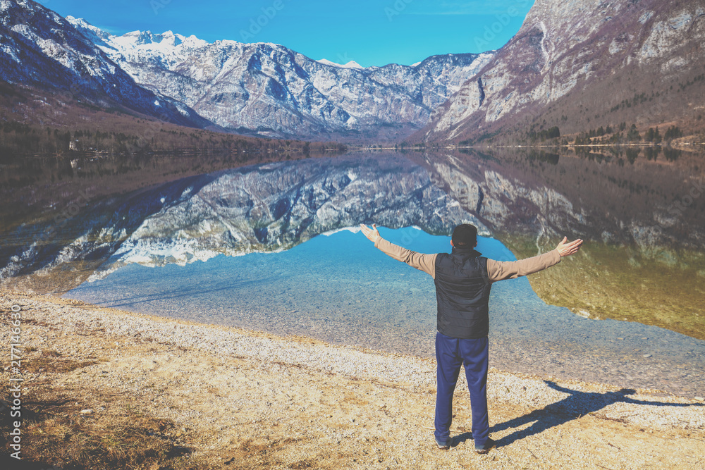 Man with hands in the air standing on the shore of the mountain lake in early spring. Bohinj lake, Slovenia, Europe