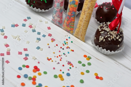 Cake pops decorated with a bow of braid. They lie on the boards, painted white. Nearby is scattered multicolored decorative topping. Visible topping in the package.