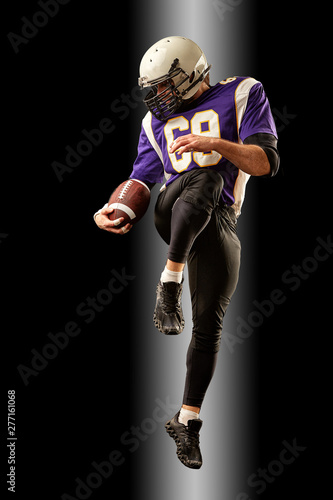 American football player holding a ball while jumping away from a strike. Black background, copy space. Violet with white sportswear.