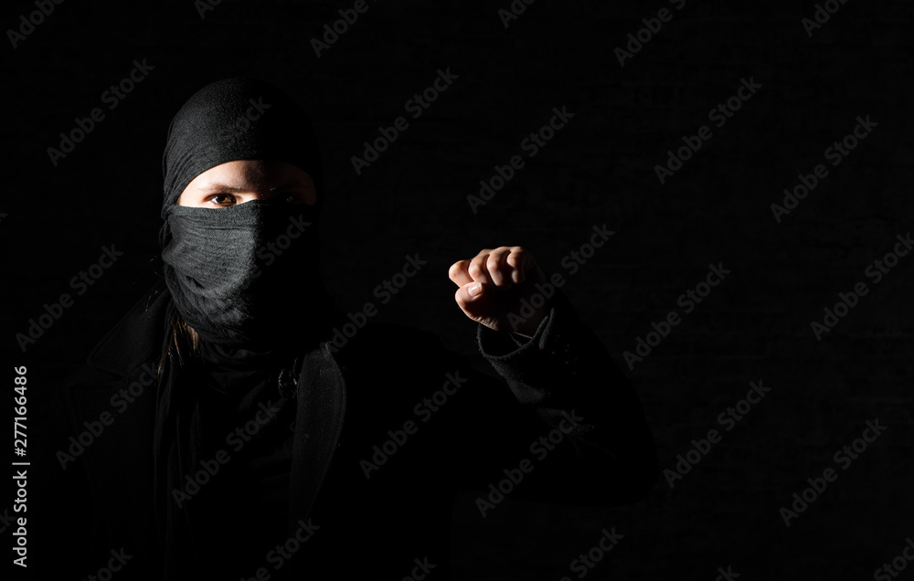 Young adult girl in black clothes with hidden face protests raising fist on black background with copy space.