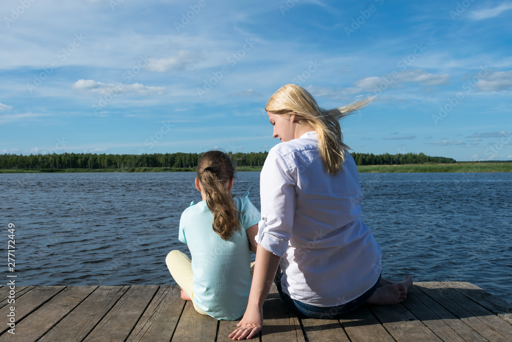 mom and daughter sit on the pier and look at the beautiful lake and nature