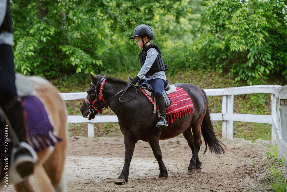 Children with helmets and protective vests on riding pony horses at sunny day on ranch.