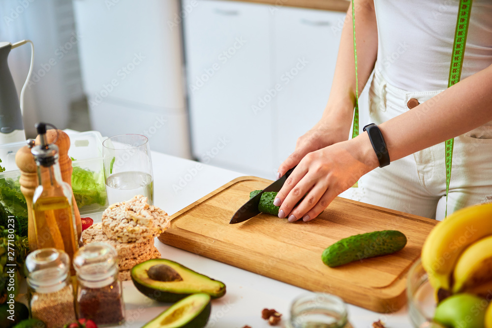 Young happy woman cutting cucumber for making salad in the beautiful kitchen with green fresh ingredients indoors. Healthy food and Dieting concept. Loosing Weight