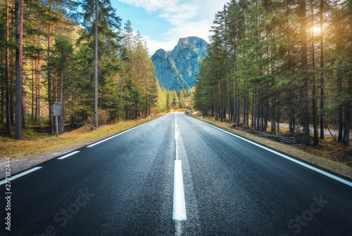 Road in summer forest at sunset in Italy. Beautiful mountain roadway, trees with green foliage and sunlight. Landscape with empty asphalt road through woodland, blue sky, high rocks. Travel in Europe