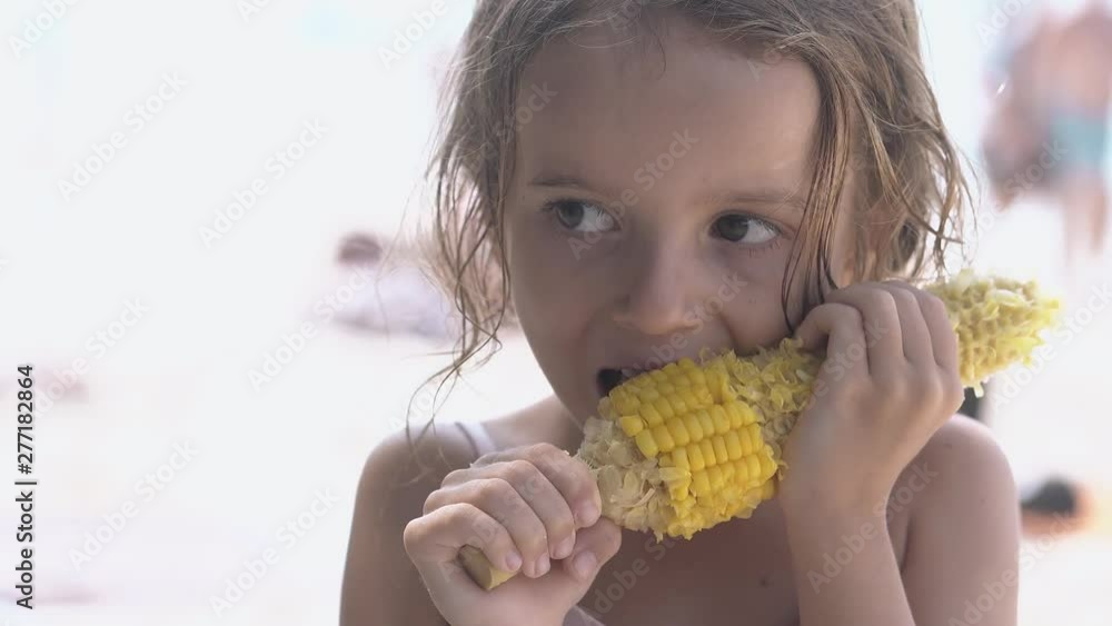 cute little girl with big brown eyes stands on beach and gnaws big corncob on hot day close view cute little girl with big brown eyes stands on beach and gnaws big corncob on hot day close view