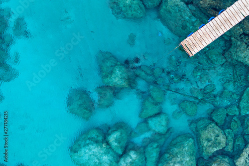 An aerial view of the beautiful Mediterranean Sea, with a wooden pier and a rocky shore, where you can see the textured underwater corals and the clean turquoise water of Protaras, Cyprus