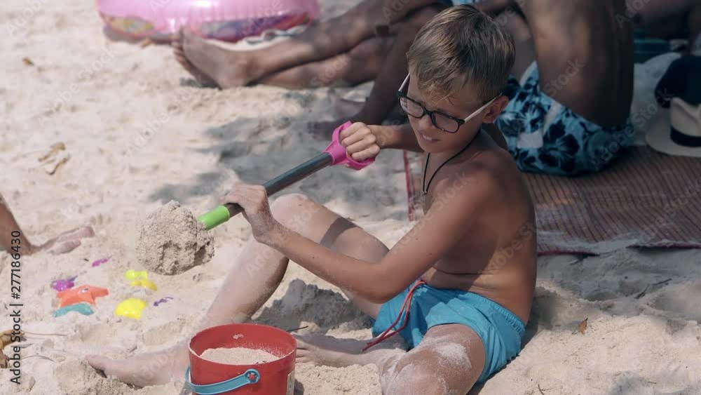 cute blond boy in glasses wearing blue swimming trunks plays with kids spade and sows sand through yellow plastic sieve cute blond boy in glasses wearing blue swimming trunks plays with kids spade and sows sand through yellow plastic sieve