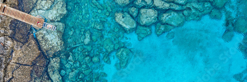 An aerial view of the beautiful Mediterranean Sea, with a wooden pier and a rocky shore, where you can see the textured underwater corals and the clean turquoise water of Protaras, Cyprus