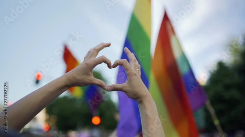Supporting hands make heart sign and wave in front of a rainbow flag flying on the sidelines of a summer gay pride parade