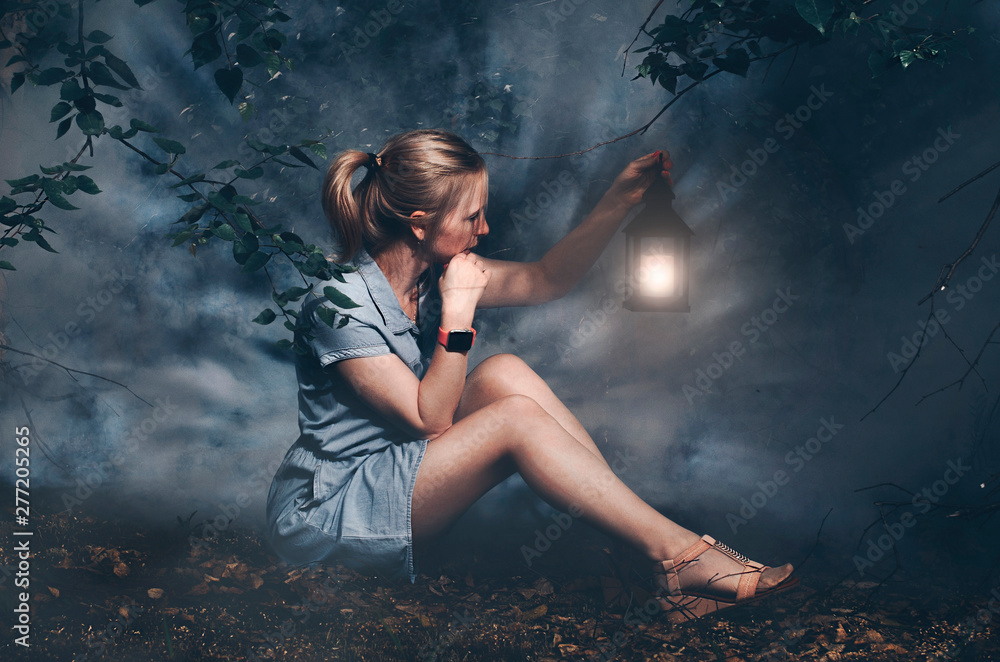 Beautiful girl in denim dress on sitting in forest with fog with lantern in hand