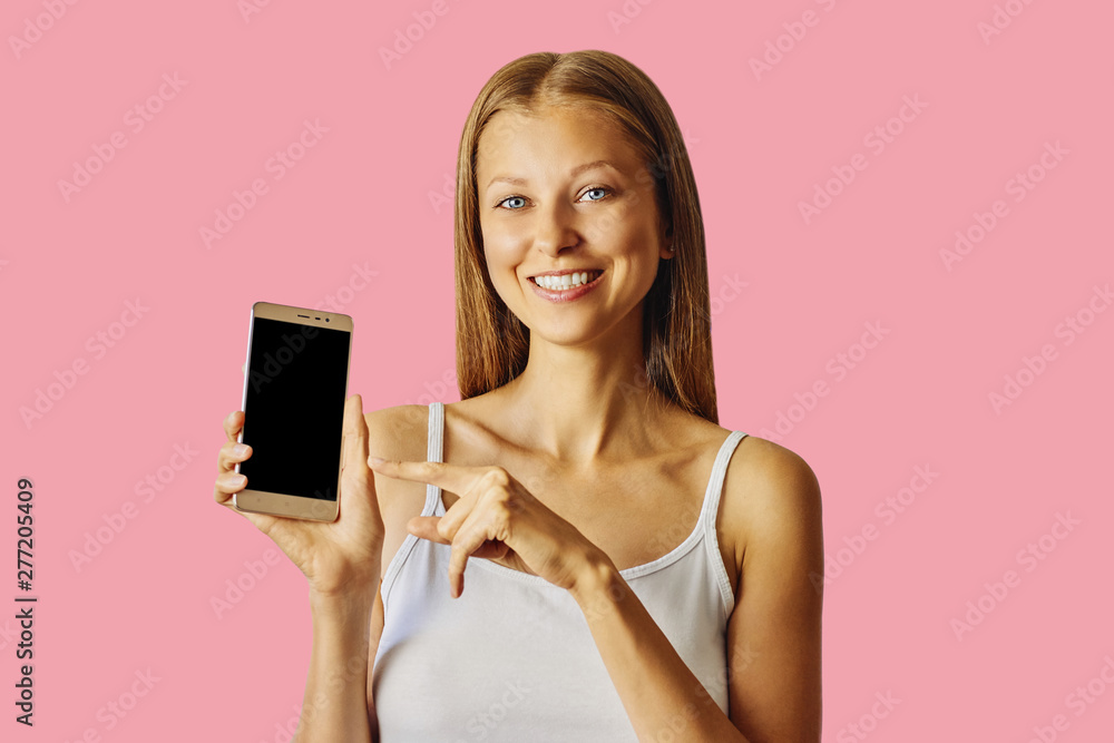 Happy smiling woman points at smartphone, isolated over pink background Happy smiling woman points at smartphone, isolated over pink background